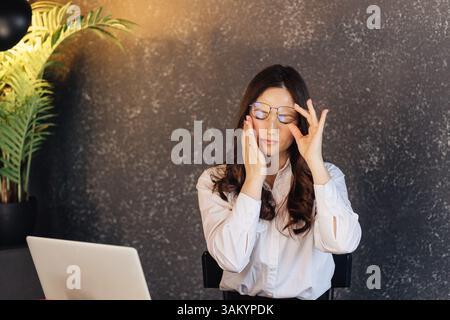 Eine Frau in einem weißen Hemd nimmt ihre Brille ab und reibt sich die Augen, was die Müdigkeit zeigt, die sie durch die Arbeit am Computer erleidet. Es symbolisiert Überlastung, digitale Augenbelastung, Stockfoto