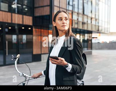 Junge selbstbewusste Geschäftsfrau mit Handy und Fahrrad im Freien stehend formelle Kleidung Junge selbstbewusste Geschäftsfrau mit Handy und Stockfoto