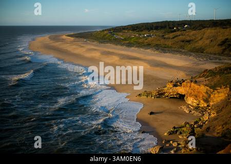 Ein sonnendurchfluteter Strand in Nazaré, Portugal, an einem klaren Tag, mit goldenem Sand, der unter einem hellblauen Himmel auf den ruhigen Atlantik trifft. Stockfoto