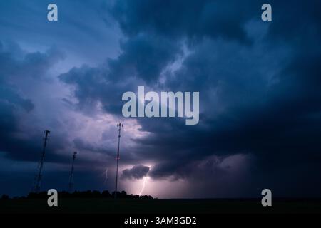 Ein dramatischer Blitzschlag während eines nächtlichen Gewitters beleuchtet dunkle Wolken über den Mobilfunktürmen in einer ländlichen Landschaft. Der intensive Himmel und der leuchtende Blitz Stockfoto