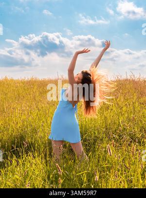 Schöne junge Frau mit gesunden langen Haaren genießt die Natur in einem grünen Grasfeld. Eine attraktive junge Frau streckt ihre Hand zum blauen Himmel. Stockfoto
