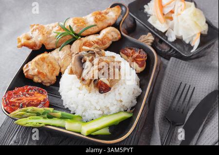 Gegrillte Hähnchenkebabs mit Reis, gebratenen Pilzen und Gemüse, serviert auf einem schwarzen Teller in gemütlicher Atmosphäre Stockfoto