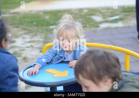 Glückliche Kinder, die sich in einem Kreisverkehr auf einem Spielplatz drehen. Die Kinder zeigen Freude und Begeisterung und genießen ihre gemeinsame Spielzeit. Grünes Gras und ein Pflaster Stockfoto
