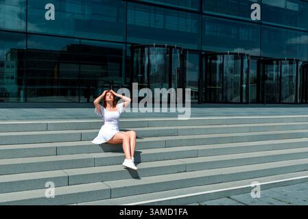 Schöne junge Frau, die auf Treppen sitzt und das helle Sonnenlicht an einem Sommertag genießt. Eine junge Frau mit weißem Kleid und Turnschuhen, die auf dem Stai sitzt Stockfoto