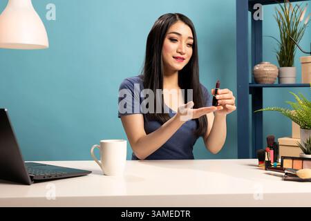 Junge Beauty-Bloggerin, die an einem Schreibtisch sitzt, Lippenstift hält und sie ihren Online-Zuschauern präsentiert. Sie zeigt sich in Richtung des Produkts, während sie ein Make-up-Tutorial-Video für ihre Livestream-Session aufnimmt. Stockfoto