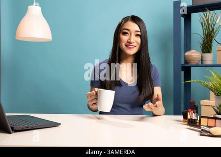Asiatische Frau lächelt und spricht ihr Online-Publikum während einer Streaming-Session im Studio mit blauem Hintergrund an. Die fröhliche Bloggerin sitzt an ihrem Schreibtisch und hält eine Kaffeetasse in der Hand, während sie in die Kamera schaut. Stockfoto