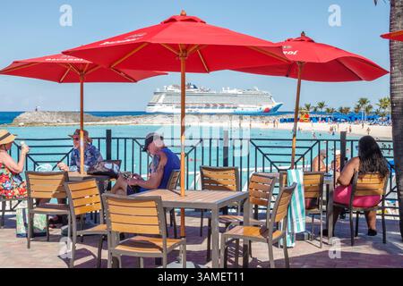 Großartiges Stirrup Cay, die Bahamas, am Strand mit roten Bacardi-Regenschirmen, Kreuzfahrtschiff vor der Küste vor Anker, Erwachsene sitzen entspannendes Resort-Café, Blick auf die Küste Stockfoto