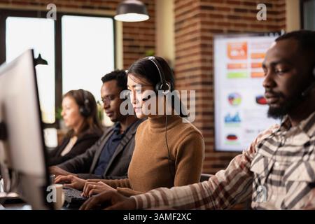 Multiethnische Callcenter-Agenten sitzen an ihren Workstations, bearbeiten Geschäftsanfragen und kommunizieren mit Remote-Kunden. Kaffee, Computer und Headsets prägen die Büroszene. Stockfoto