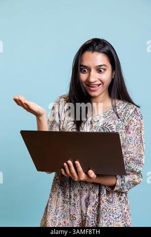 Fröhliche Person, die mit Freunden bei einer Videokonferenz tratscht, posiert vor dem Hintergrund des Studios. Glückliche, entspannte Frau, die sich mit ihren Mädchen verbündet, nehmen Sie an Online-Videogesprächen auf dem Laptop Teil. Stockfoto
