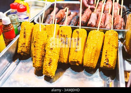 Gegrillter Mais auf Spießen, ausgestellt an einem lebhaften Street Food-Stand, mit saftigen Würstchen im Hintergrund. Perfekt für Sommerkonzepte Stockfoto