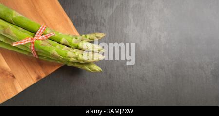 Grüne Spargelspeere aus nächster Nähe. Ein Haufen frischer roher grüner Spargel auf einem hölzernen Schneidebrett auf grauem Hintergrund mit Kopierraum-Banner von oben. Stockfoto