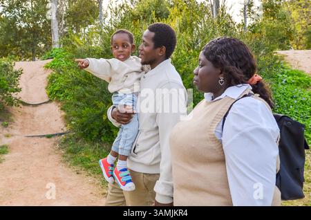 Lächelnde afroamerikanische Familie macht einen Spaziergang im Park, während ihr Kind auf etwas in der Ferne zeigt Stockfoto