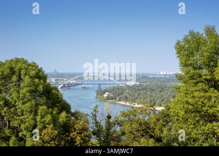 Ein malerischer Blick durch üppige grüne Bäume fängt den Fluss Dnipro ein, der unter der Podilsko-Voskresenskyi-Brücke fließt, in Richtung Kiews Stadtlandschaft unter einem cl Stockfoto