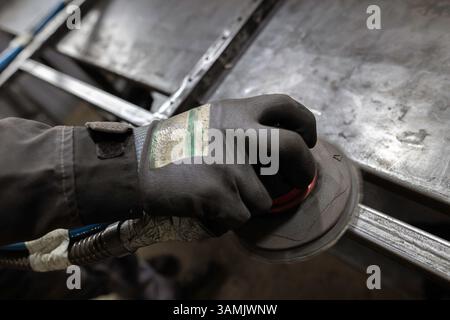 Die Handschuhe eines Arbeitnehmers verwenden einen Schleifmaschinen, um die Fugen eines Metallrahmens auf einer Werkbank in einer Werkstatt zu glätten. Stockfoto