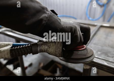 Die Handschuhe eines Arbeitnehmers verwenden einen Schleifmaschinen, um die Fugen eines Metallrahmens auf einer Werkbank in einer Werkstatt zu glätten. Stockfoto