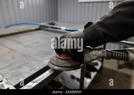 Die Handschuhe eines Arbeitnehmers verwenden einen Schleifmaschinen, um die Fugen eines Metallrahmens auf einer Werkbank in einer Werkstatt zu glätten. Stockfoto