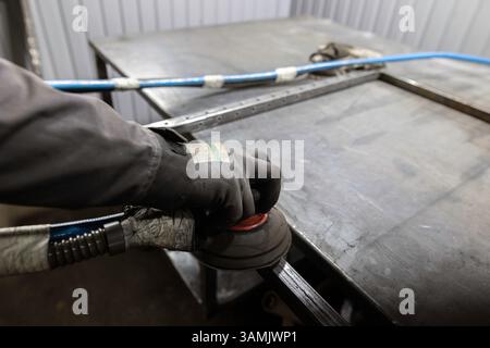 Die Handschuhe eines Arbeitnehmers verwenden einen Schleifmaschinen, um die Fugen eines Metallrahmens auf einer Werkbank in einer Werkstatt zu glätten. Stockfoto