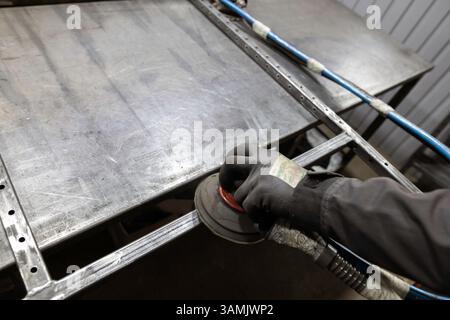 Die Handschuhe eines Arbeitnehmers verwenden einen Schleifmaschinen, um die Fugen eines Metallrahmens auf einer Werkbank in einer Werkstatt zu glätten. Stockfoto