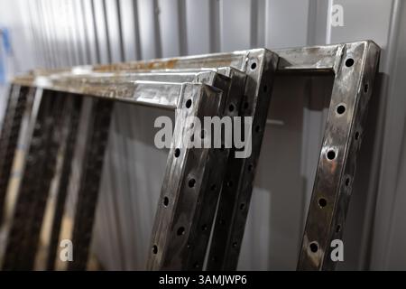 Mehrere geschweißte Metallrahmen mit gebohrten Löchern werden in einer Werkstattumgebung gegen eine gewellte Metallwand gestapelt. Stockfoto