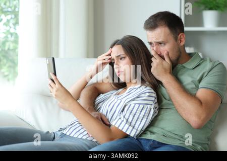 Besorgtes Paar, das Online-Informationen auf dem Mobiltelefon überprüft und zu Hause auf einer Couch sitzt Stockfoto