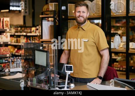 Ein lächelnder Kassierer mit Bart, der hinter der Kasse des Lebensmittelgeschäfts steht, trägt ein braunes Hemd und ein Personalabzeichen, um Kunden zu begrüßen und ihnen zu helfen Stockfoto