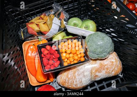 Einkaufswagen gefüllt mit verschiedenen frischen Lebensmitteln, einschließlich Paprika, Himbeeren, Brokkoli, Äpfeln und Brot umgeben von verpackten Nudeln und Tomaten Stockfoto
