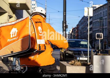 Posen, Polen - 8. April 2025: Rückansicht eines Pyszne.pl-Kuriers auf einem Fahrrad, der eine leuchtend orange Jacke und einen isolierten Lieferrucksack trägt Stockfoto