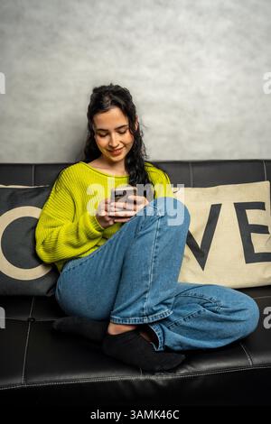 Die junge Frau benutzt ihr Smartphone, während sie sich zu Hause auf einem bequemen Sofa entspannt Stockfoto