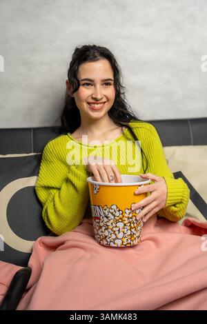 Lächelnde junge Frau, die zu Hause einen Filmabend genießt und Popcorn aus einem großen Eimer isst Stockfoto