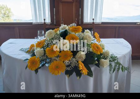 Ein elegantes, florales Herzstück aus gelben Gerbera-Gänseblümchen und weißen Rosen ziert einen Tisch mit einer weißen Tischdecke in Italien, geeignet für Hochzeitsempfänge oder besondere Veranstaltungen. Hochwertige Fotos Stockfoto