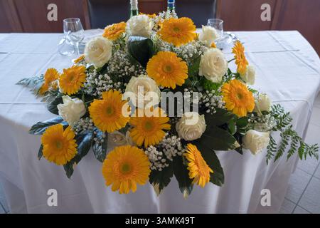 Ein elegantes, florales Herzstück aus gelben Gerbera-Gänseblümchen und weißen Rosen ziert einen Tisch mit einer weißen Tischdecke in Italien, geeignet für Hochzeitsempfänge oder besondere Veranstaltungen. Hochwertige Fotos Stockfoto