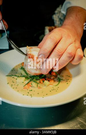 Ein Mann legt Essen auf einen Teller. Der Teller hat einen weißen Hintergrund und das Essen ist eine Mischung aus Fleisch und Gemüse. Der Mann benutzt eine Gabel, um den Fo zu vertreiben Stockfoto