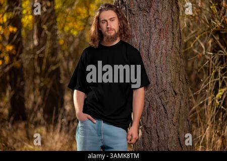 Modell eines schwarzen übergroßen T-Shirts auf einem Mann mit lockigen Haaren, Bart, mit Hand in der Tasche, draußen, auf dem Hintergrund von Bäumen, gelben Blättern. Vorlage Stockfoto