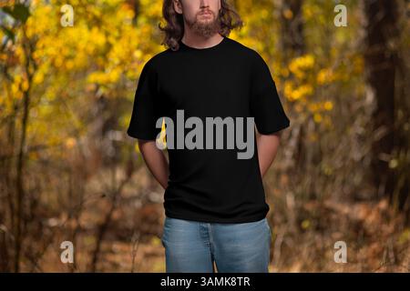 Modell eines schwarzen übergroßen T-Shirts auf einem Mann mit langen Haaren, einem Bart, einer Vorderansicht, draußen, auf dem Hintergrund von Bäumen, gelben Blättern. Vorlage eines Stockfoto