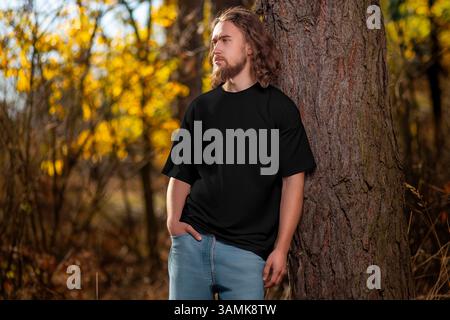 Übergroßes schwarzes T-Shirt-Modell auf einem Mann mit lockigen Haaren, Bart, stehend mit dem Rücken zu einem Baum, draußen, im Hintergrund der Natur. Männliche Streetwea Stockfoto
