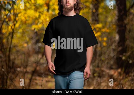 Übergroße schwarze T-Shirt-Vorlage auf einem bärtigen Mann mit langen lockigen Haaren, der auf einem Naturhintergrund läuft, Vorderansicht. Hemd-Mockup für einen Kerl, draußen. Pr Stockfoto