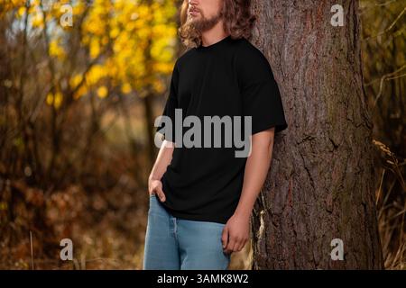 Übergroße schwarze T-Shirt-Vorlage auf einem bärtigen Mann mit lockigen Haaren, zurück zu einem Baum, Seitenansicht, auf dem Hintergrund der herbstlichen Natur, draußen. Männliche Fashio Stockfoto