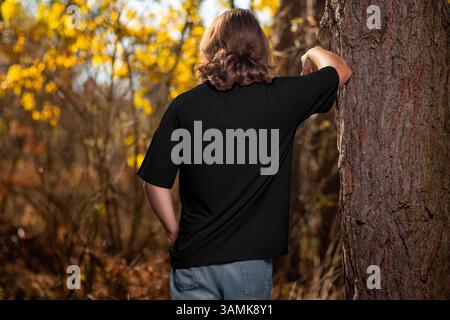 Vorlage eines schwarzen übergroßen T-Shirts auf einem Mann mit langen lockigen Haaren, auf einem Baum gelehnt, Rückansicht, draußen. Mockup eines Hemdes an einem Kerl im Hinterkopf Stockfoto