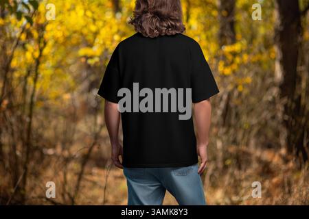 Übergroßes schwarzes T-Shirt Mockup auf einem Mann mit langen Haaren, Rückansicht, auf dem Hintergrund der herbstlichen Natur, draußen. Schablonenhemd für Herren, zum Design Stockfoto