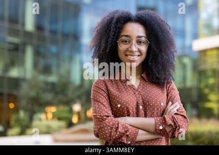 Eine selbstbewusste junge Frau mit lockigem Haar und Brille lächelt in die Kamera. Sie steht draußen mit überkreuzten Armen, ein modernes Gebäude im Hintergrund. Stockfoto