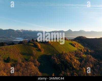 Blick aus der Vogelperspektive auf die malerische katholische Kirche St. Primus und Felicianer auf einem ruhigen Hügel umgeben von Herbstwald und Bergen, Jamnik, Kranj, Stockfoto