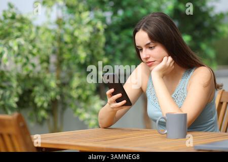 Frau, die ein Telefon benutzt, sitzt auf einer Terrasse zu Hause an einem sonnigen Tag Stockfoto