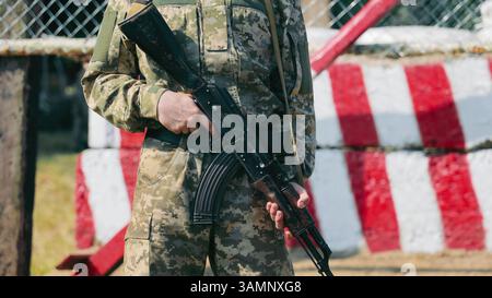 Soldat mit Gewehr vor der Roten und Weißen Barriere. Ein Soldat in Tarnuniform hält ein Gewehr, während er an einem Kontrollpunkt bereit steht. Stockfoto