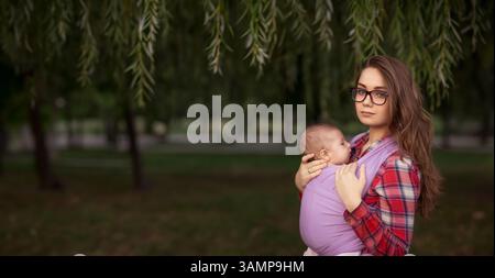 Junge Mutter mit Baby in Schlinge unter Weidenbaum im Park. Friedliche Bindung und Mutterschaft Stockfoto