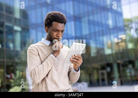 Ein nachdenklicher Geschäftsmann untersucht sein Tablet und scheint Informationen mit einer Hand am Kinn zu erwägen, draußen in der Nähe eines modernen Gebäudes. Stockfoto