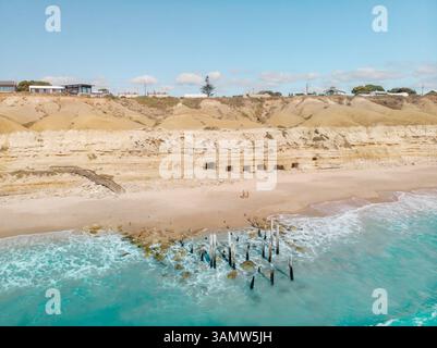 Blick aus der Vogelperspektive auf den Strand von Port Willunga mit Holzpfählen im Wasser und zwei Personen, die am Strand stehen, mit Höhlen in der Klippenwand dahinter, Süden Stockfoto