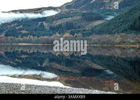Niedriger Nebel, der sich am Berg anschmiegt, und die Reflexionen herbstlicher Wälder am Ufer, im ruhigen Wasser von Loch Goilhead. Stockfoto