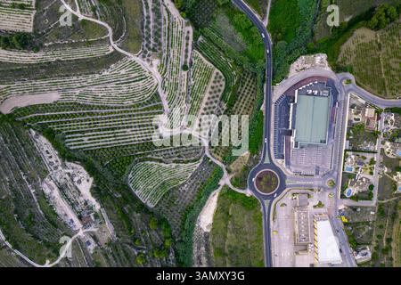 Aus der Vogelperspektive auf eine Serpentinenstraße und einen modernen Gebäudekomplex, in der Nähe grüner landwirtschaftlicher Felder, Zona Encinas, Cumbre del Sol, Alicante, Spanien. Stockfoto