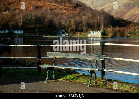 Eine verwitterte Bank aus Holz und Gusseisen am Stronachlachar Pier mit dem Dorf dahinter über dem Wasser von Loch Katrine. Stockfoto