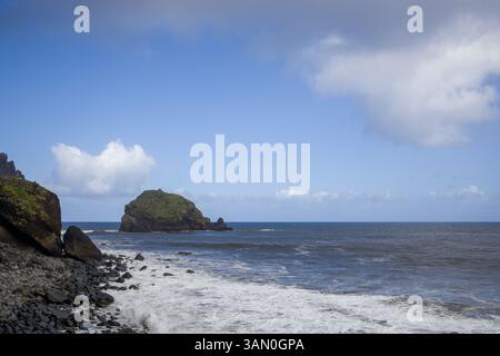 : Das Bild fängt Madeiras zerklüftete Küste ein, mit massiven Felsbrocken und Wellen, die unter einem leuchtend blauen Himmel mit Wolken krachen, und hebt die hervor Stockfoto
